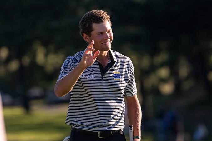 Michael Brennan, in a striped polo, smiles and raises his hand to the crowd